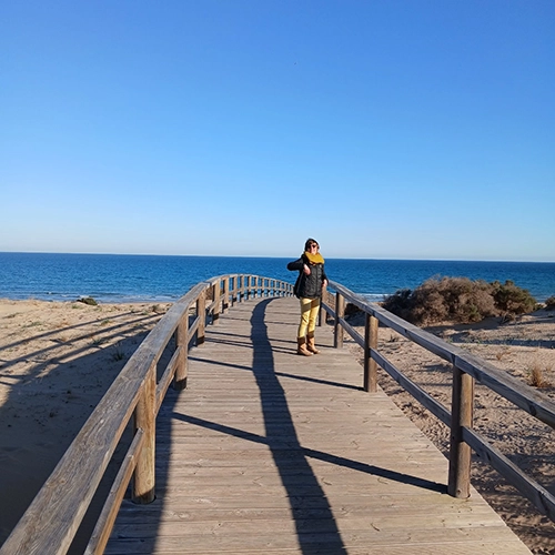 En medio de una playa una mujer sonríe de pie en medio de un camino de madera